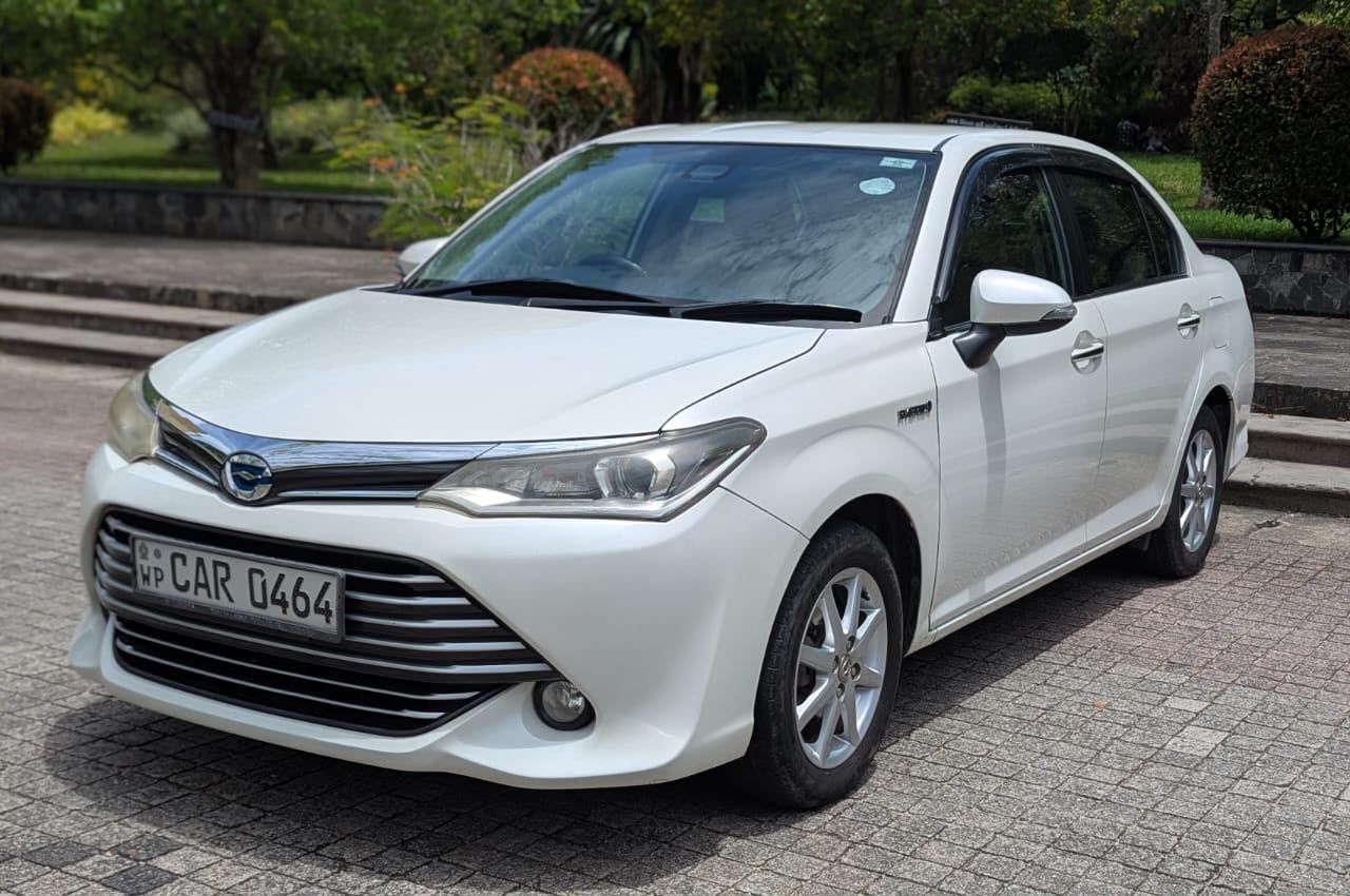 Ceylon Now Economy vehicle, a white Toyota Axio Hybrid sedan, parked on paving beside a garden in Sri Lanka